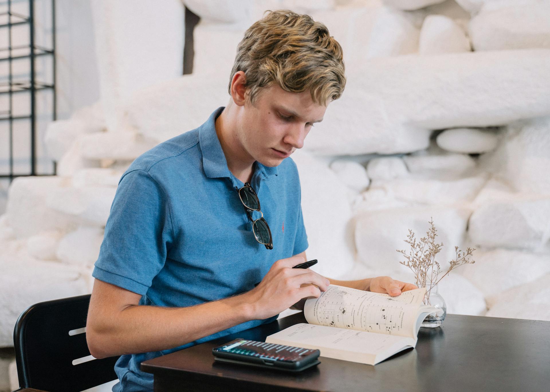 A young man in a blue polo shirt studying with a book and calculator on a table indoors.