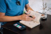 一名身穿蓝色衬衫的男子在室内桌旁用智能手机和书籍学习