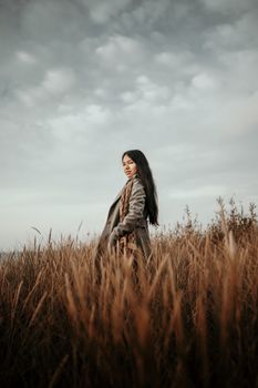 A woman stands in a field during fall, surrounded by tall grasses under a cloudy sky.
