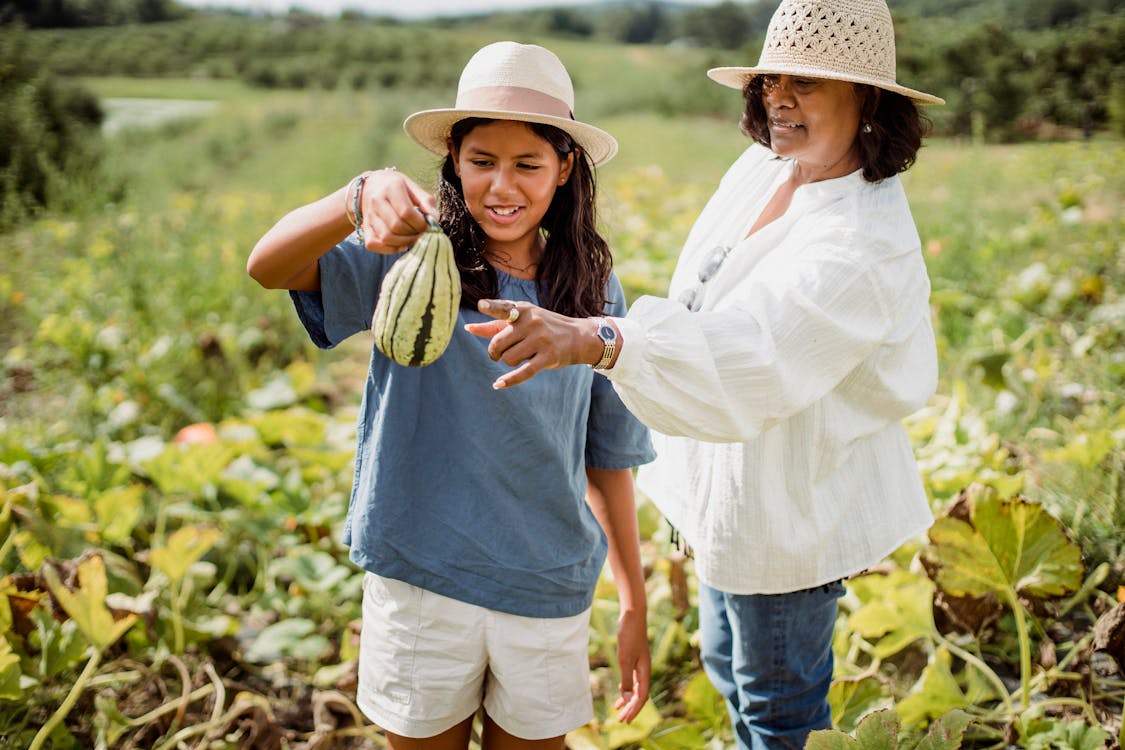 Smiling Latin American young girl holding unripe pumpkin while standing with mother in agricultural field
