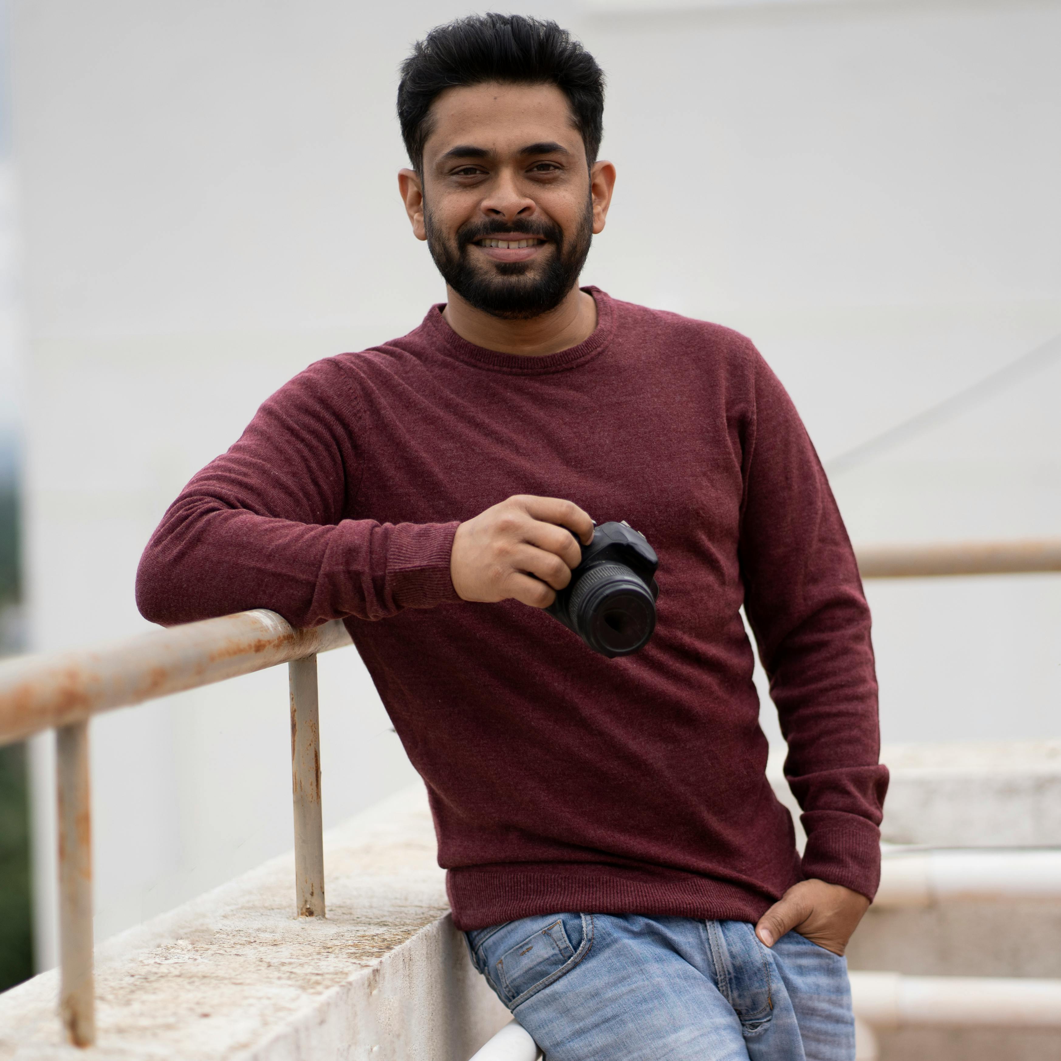 Indian man in casual attire holding a camera, smiling outdoors.