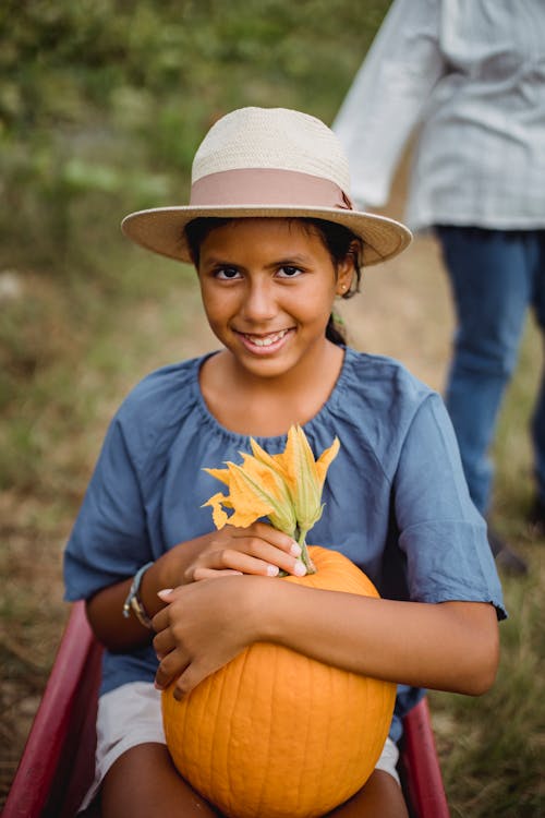 Young Latin American teen girl in hat holding pumpkin while sitting in cart and looking at camera
