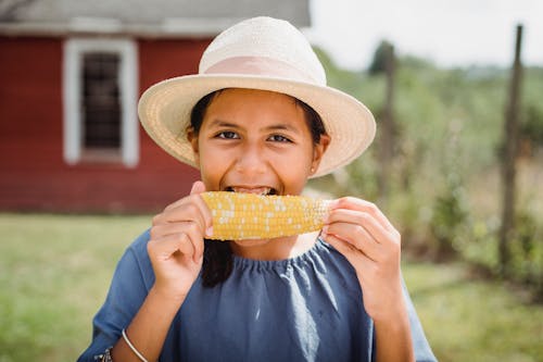 Cheerful Hispanic girl in hat biting tasty appetizing corn while spending summer day in yard