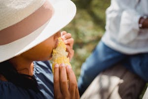 From above of ethnic anonymous teen girl in straw hat enjoying sweet corn while having lunch in yard