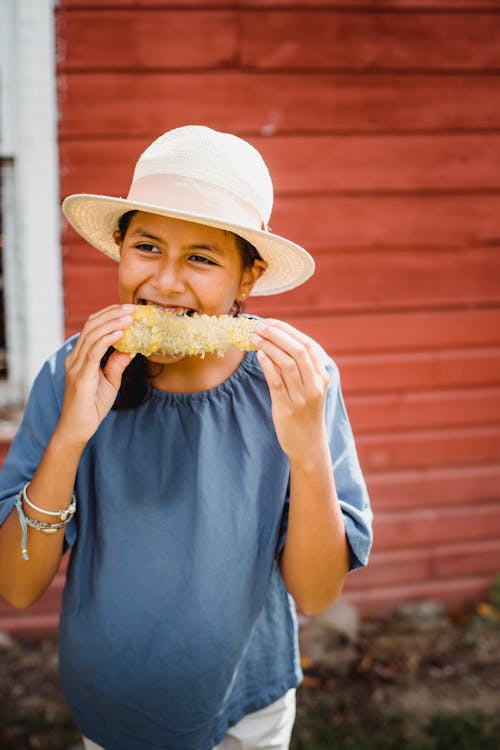 Smiling ethnic girl eating corn against wooden house