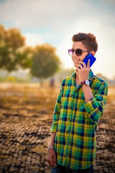 Teenage boy in sunglasses and plaid shirt talks on phone in a sunny outdoor setting.