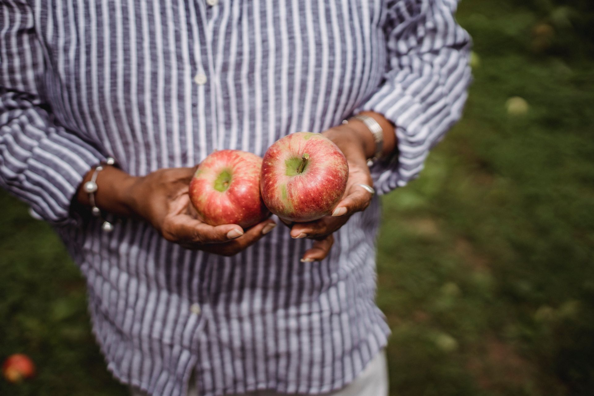 Anonymous ethnic female wearing striped shirt standing in orchard with apples on blurred background during harvest season on summer day