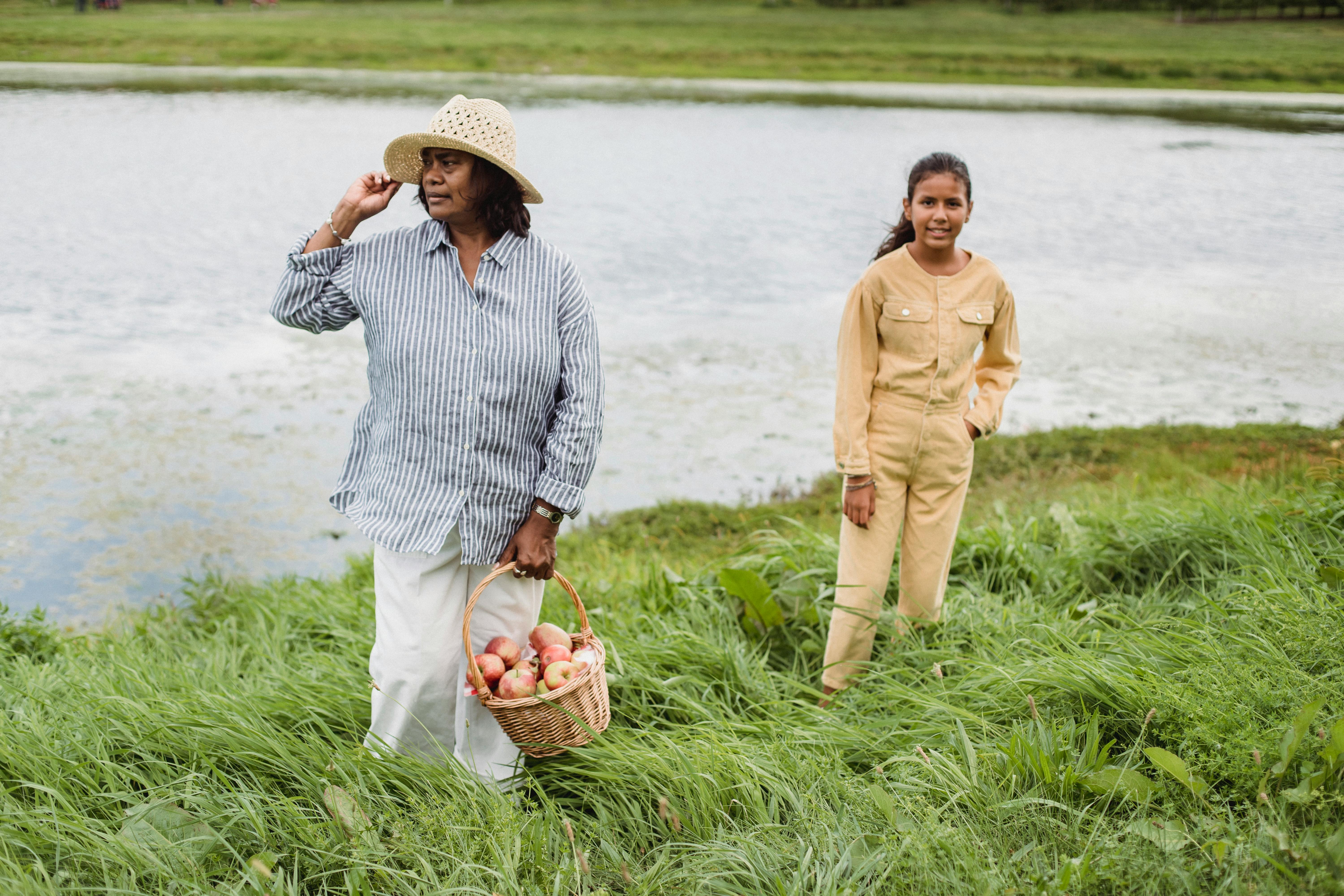 A woman and a young girl enjoy harvesting apples outdoors near a serene pond.