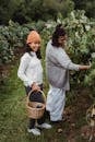 Full length of cheerful ethnic females picking up black grapes in wicker basket in countryside in soft daylight