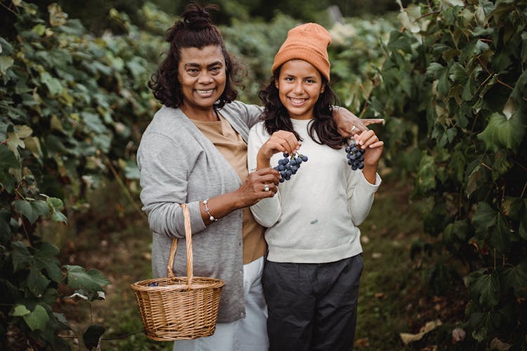 Happy Ethnic Woman Embracing Teenager With Bundles Of Ripe Grapes