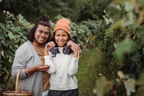 Glad ethnic woman embracing teenager with bundles of ripe grapes in vineyard while looking at camera