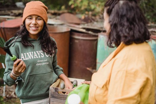 Happy ethnic girl with gardening tool and box looking at camera near crop unrecognizable mother on farm