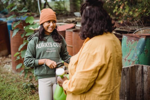 Cheerful ethnic teenager speaking with anonymous gardener with watering can while looking away on farm in daylight