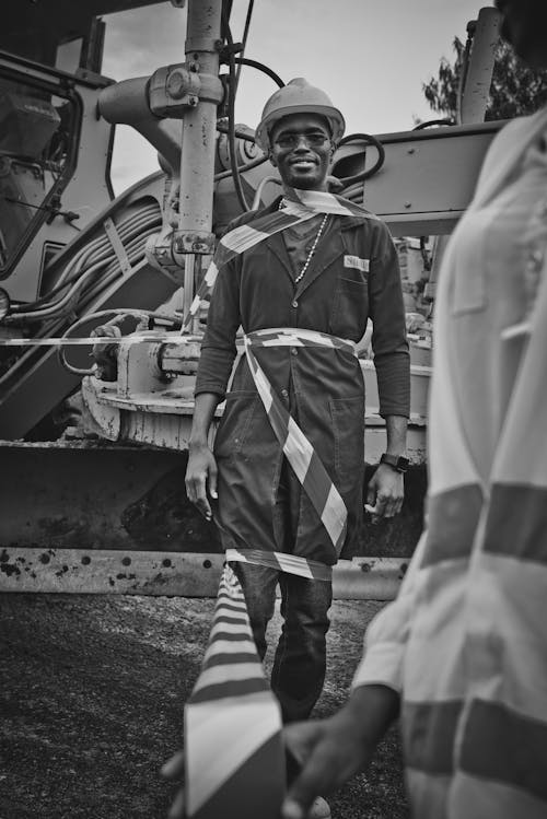 Black and white positive male African American mechanic in uniform wrapped in warning tape standing near excavator