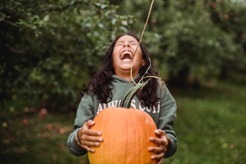 Laughing ethnic teenager with closed eyes standing with fresh pumpkin on pathway near trees on farm