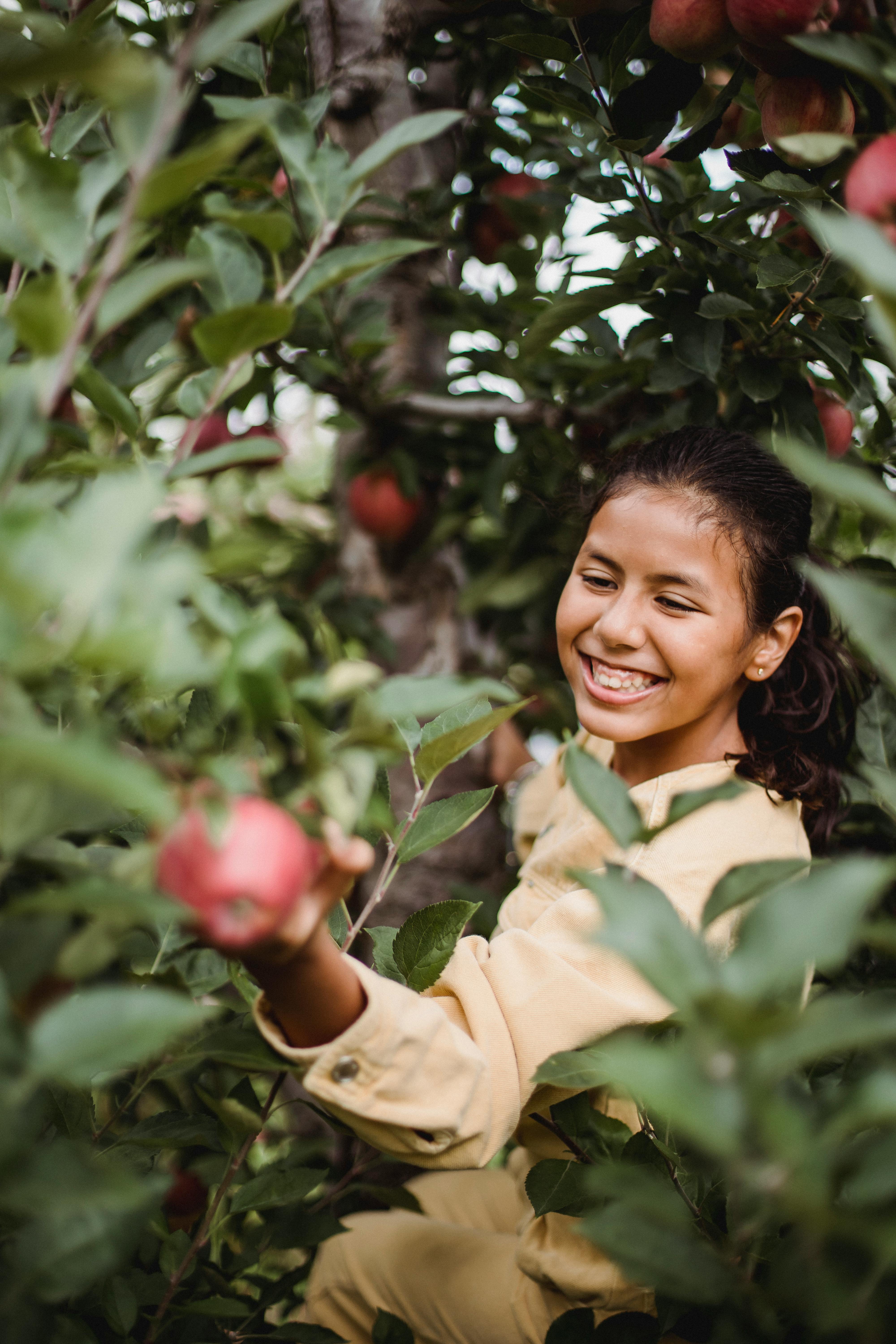 smiling ethnic gardener with apple among tree foliage