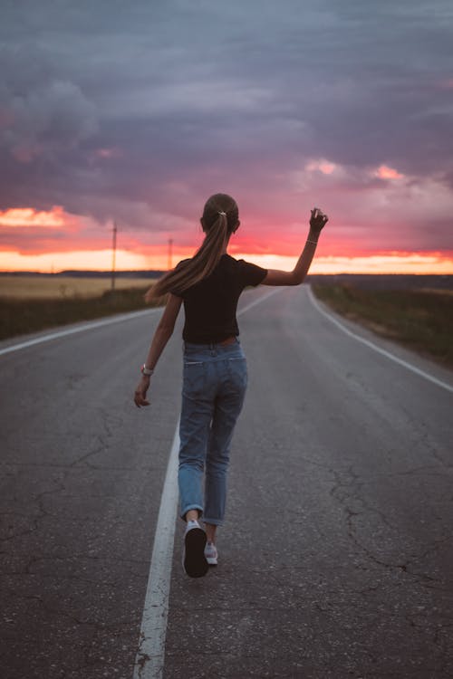 Back view of confident teenage girl with long ponytail waving hand walking confidently on rural roadway in sunset