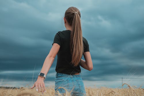Low angle of teen girl touching spikelets strolling in golden field under stormy gray sky