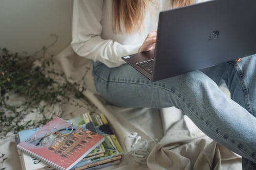 Free Crop anonymous woman in denim working on laptop sitting on bed with stack of books and notepads Stock Photo