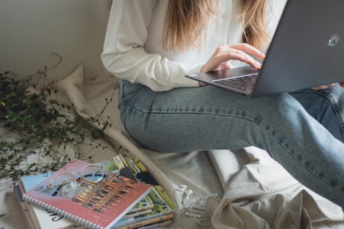 Free Faceless casual woman studying on laptop sitting with pile of textbooks and earphones at home Stock Photo