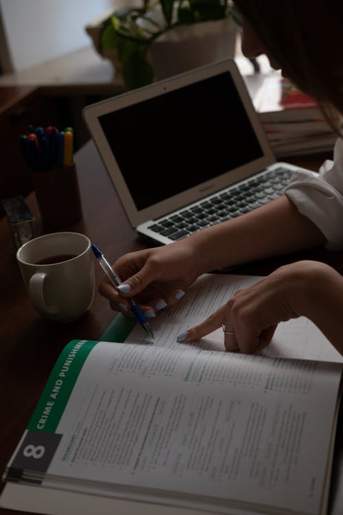 Crop side view of female student at desk with laptop doing tasks in textbook