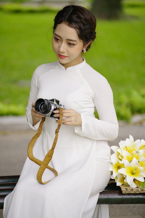 Optimistic Asian female photographer in stylish white outfit sitting near flowers with photo camera in hands in park with green lawn