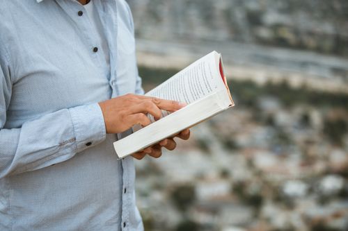 Free A person in a blue shirt reading a book, standing outdoors with a blurred cityscape in the background. Stock Photo