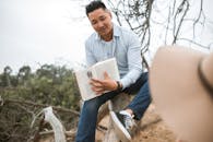 A Man in a Long Sleeved Shirt Showing a Book while Sitting on a Tree Trunk