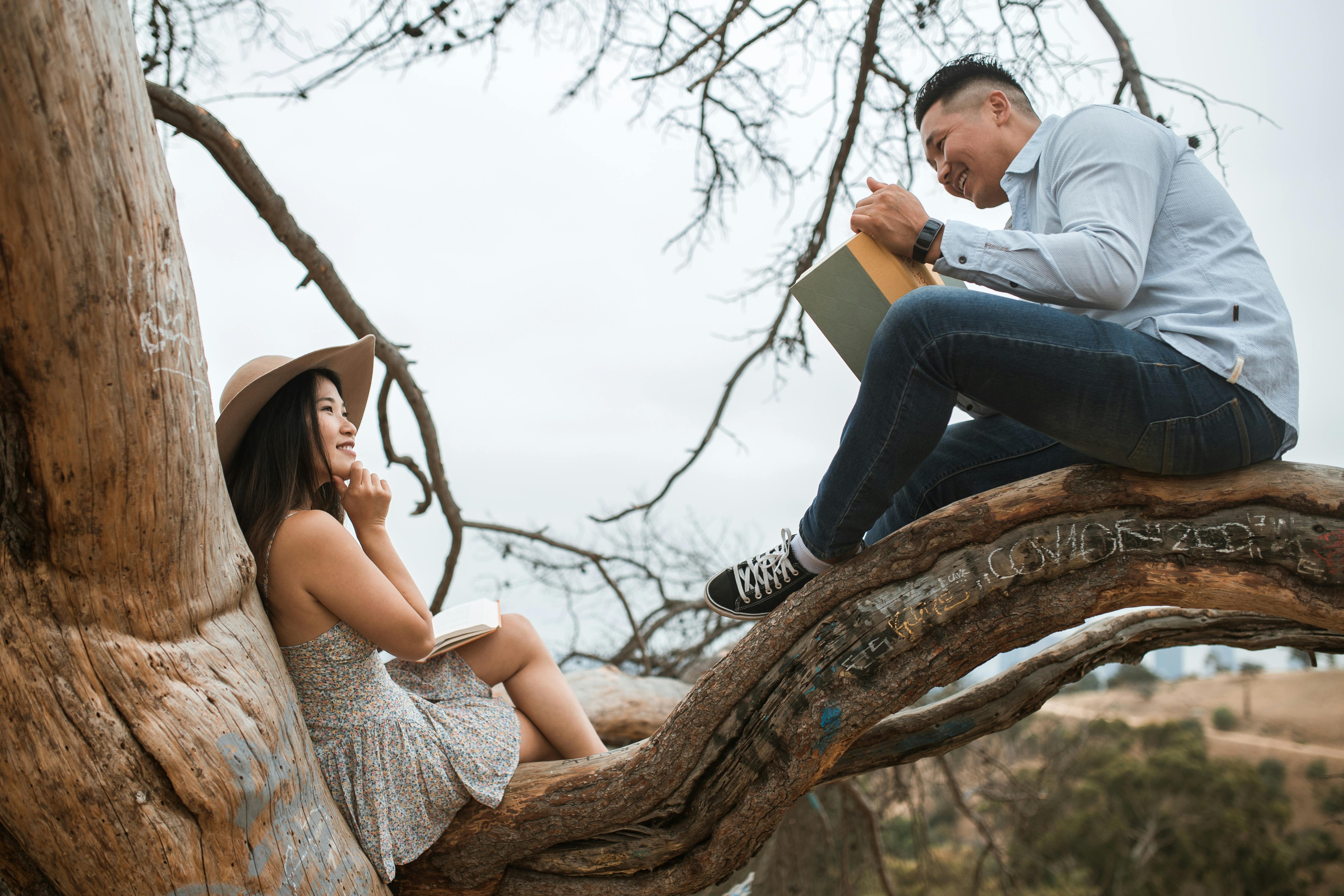 A couple enjoys reading together while sitting on a large tree branch in a serene outdoor environment.