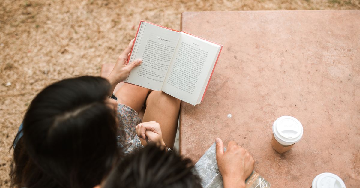 A person reading a physical book on a park bench with a coffee cup, no phone in sight