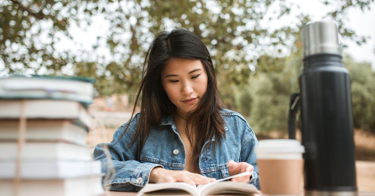 A student sitting on a park bench reading a dog-eared book with a coffee cup, surrounded by autumn leaves