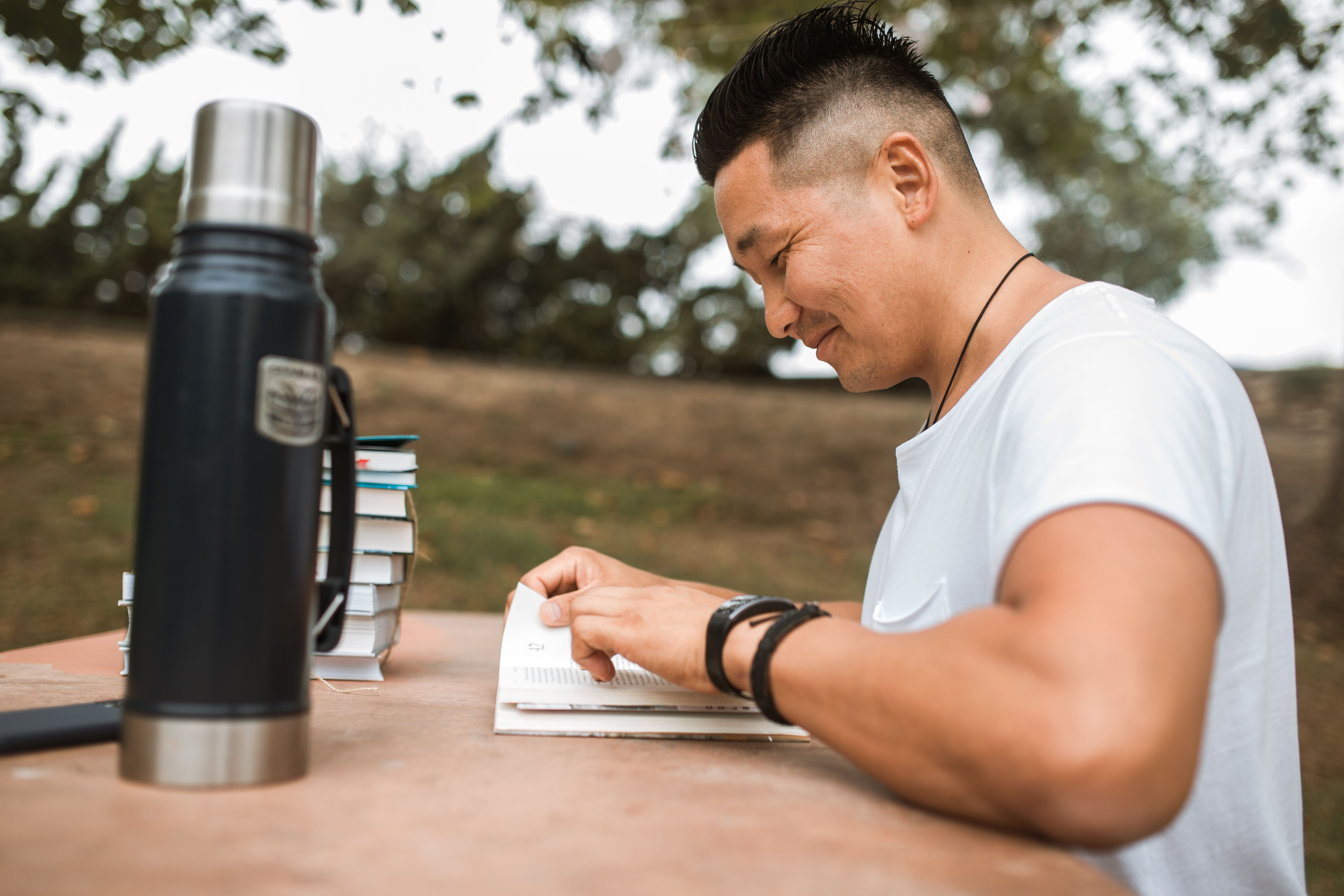 Man enjoying reading at an outdoor table with books and a thermos. Perfect study or relaxation scene.