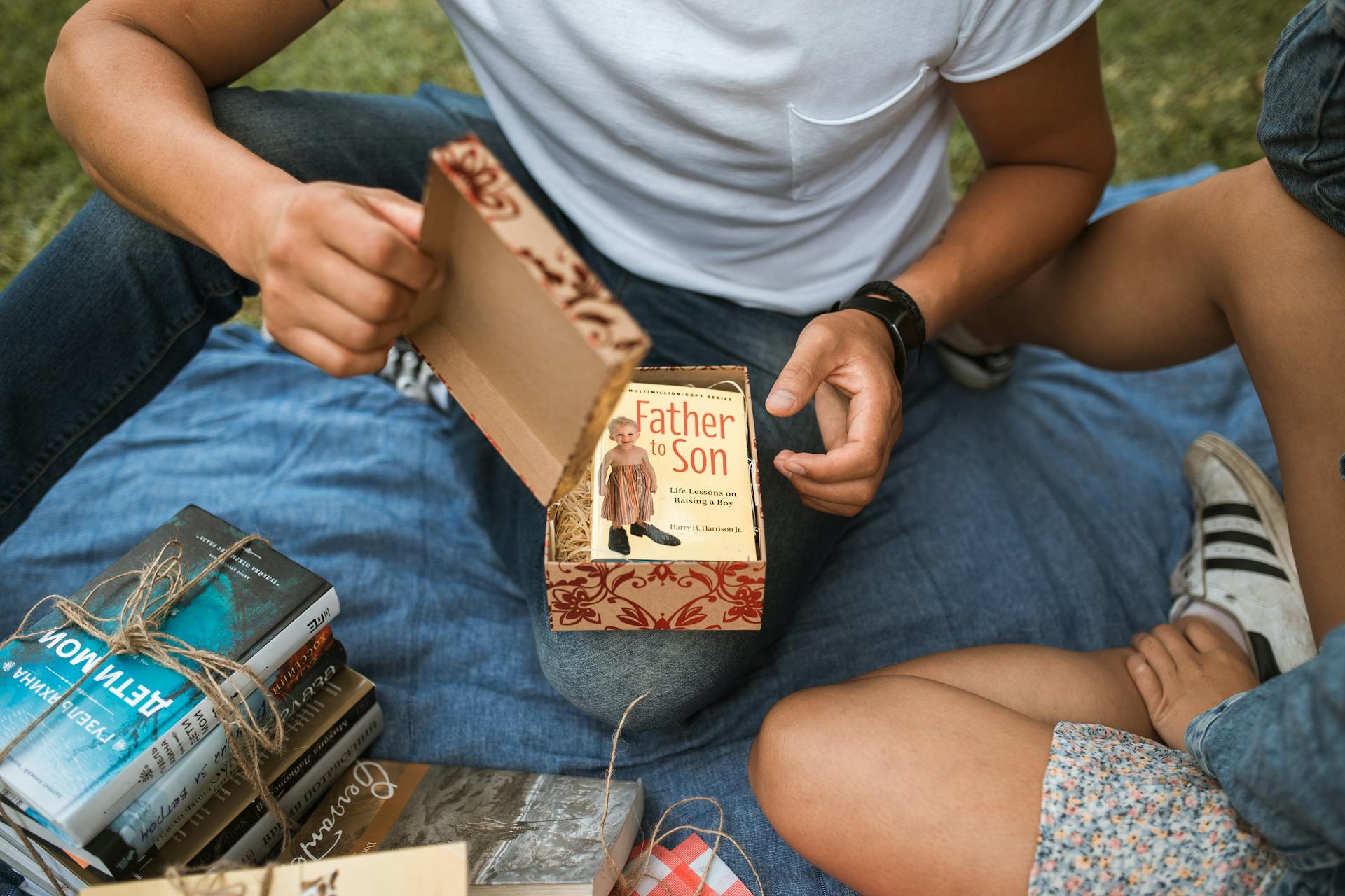 A person in a white shirt opens a gift box containing the book 'Father to Son' during a picnic.