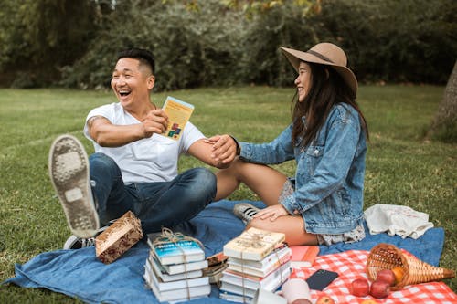 Free Man in White T-Shirt Holding a Book and Sitting Beside Woman in Hat Stock Photo