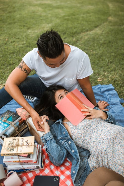Free Woman in Floral Dress Lying Beside Man in White T-Shirt and Holding Red Book Stock Photo
