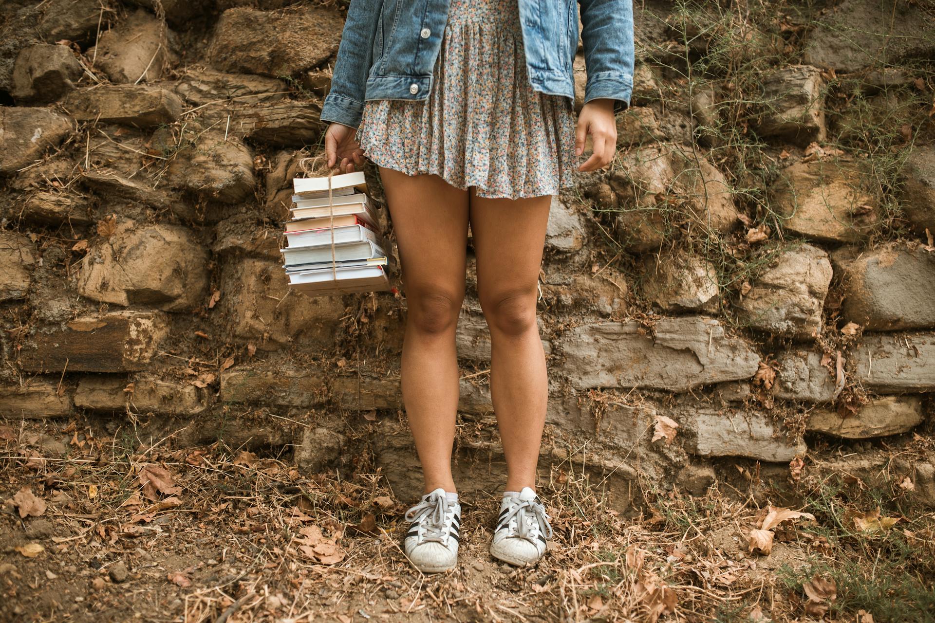 Person in Denim Jacket and Floral Mini Dress Standing on Dirt Ground Holding a Pile of Books