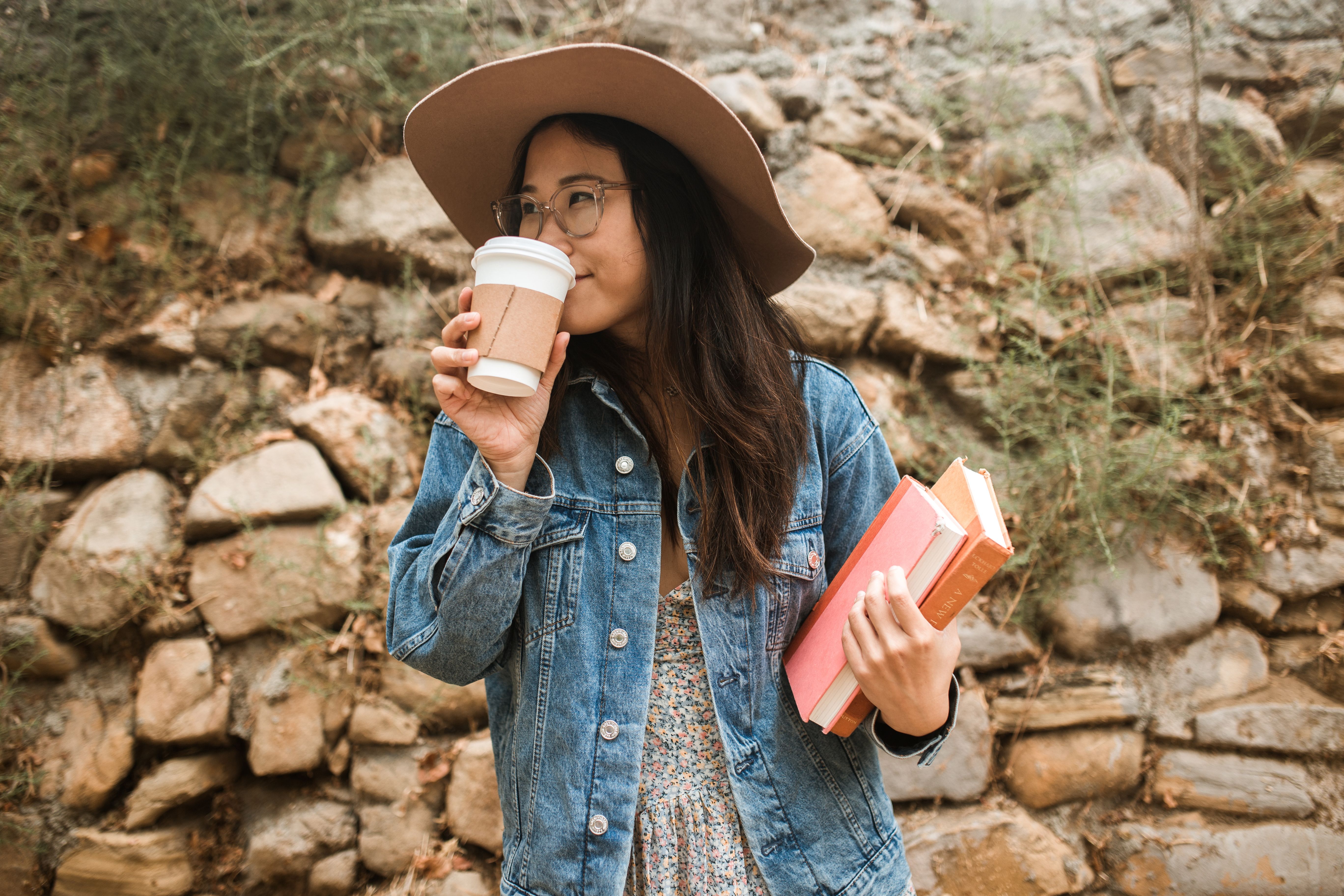 Casual woman in denim sipping coffee and holding books against a stone wall background.