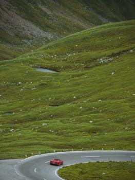 From above of red sports car driving on empty asphalt road among hills covered with green grass in daytime