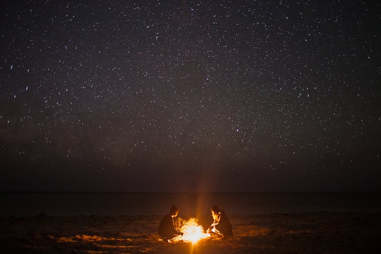 Unrecognizable Couple Near Bonfire On Coast At Night Sky