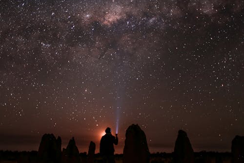 Back view silhouette of anonymous person shining flashlight at sky with glowing stars while sitting in nature at night time