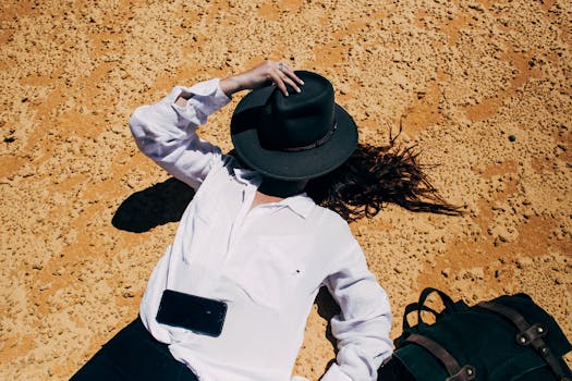 Woman in white blouse and hat relaxing on sandy beach with smartphone on chest.