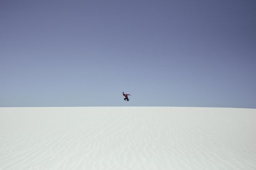 Free Silhouette of a person energetically jumping on a snowy hill under a bright blue sky. Stock Photo