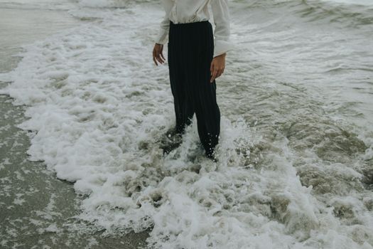 一个人站在海滩上，海浪拍打着他的黑色裤子，营造出宁静的沿海景象