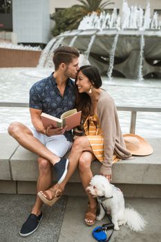 一对幸福的夫妇和他们的狗坐在喷泉边，在户外微笑着阅读