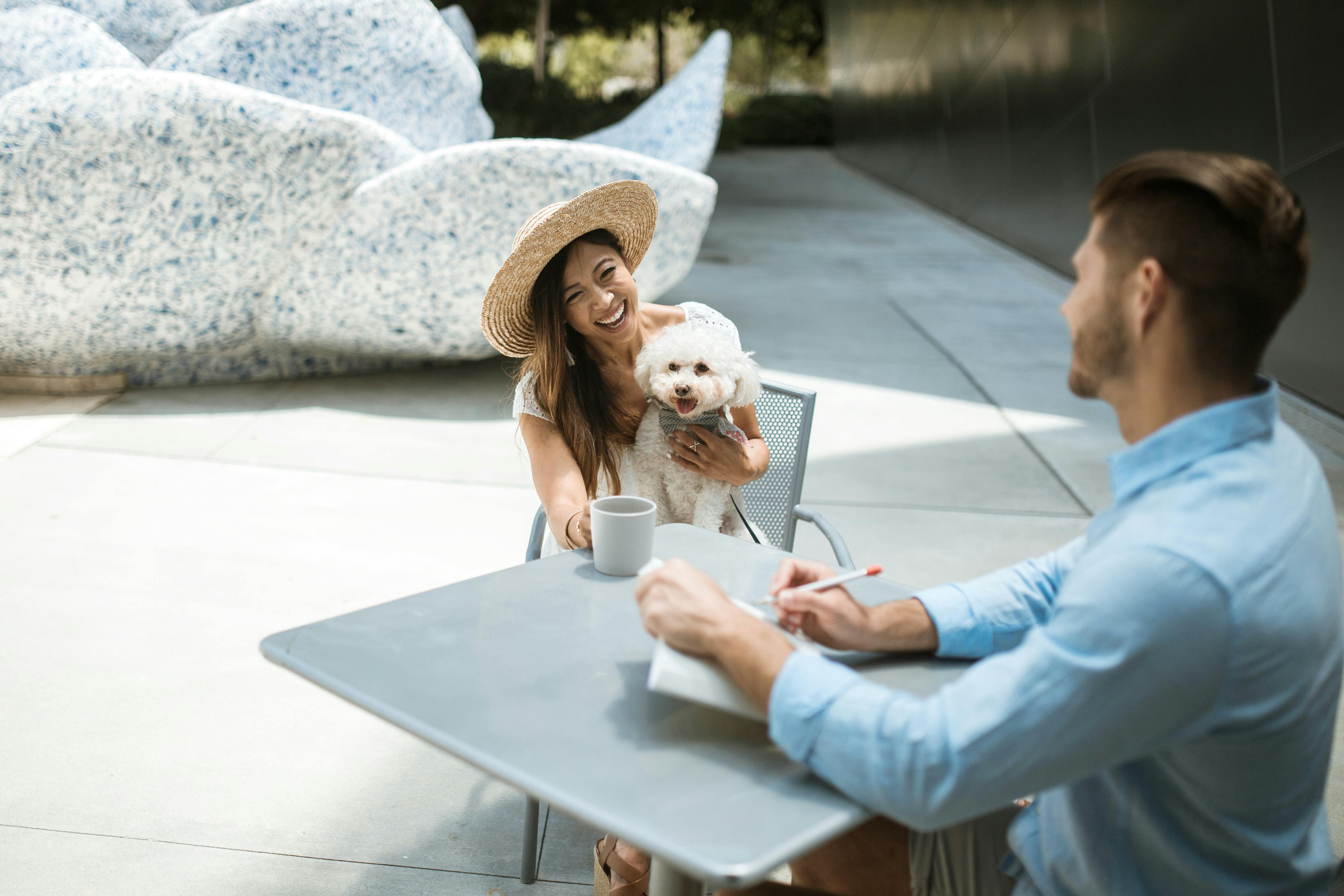 阳光明媚的户外咖啡馆里，一个戴着草帽的女人抱着她的狗，进行了一次友好的会面