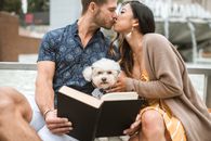 一对夫妇在城市喷泉旁与他们的贵狗一起读书时，分享了一个吻