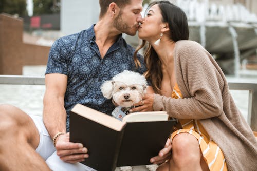 A couple shares a kiss while reading a book with their poodle by a city fountain.