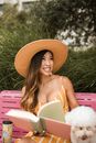 一位穿着夏装、戴着帽子的开朗女人，喜欢和她的蓬松同伴一起在户外的长凳上阅读