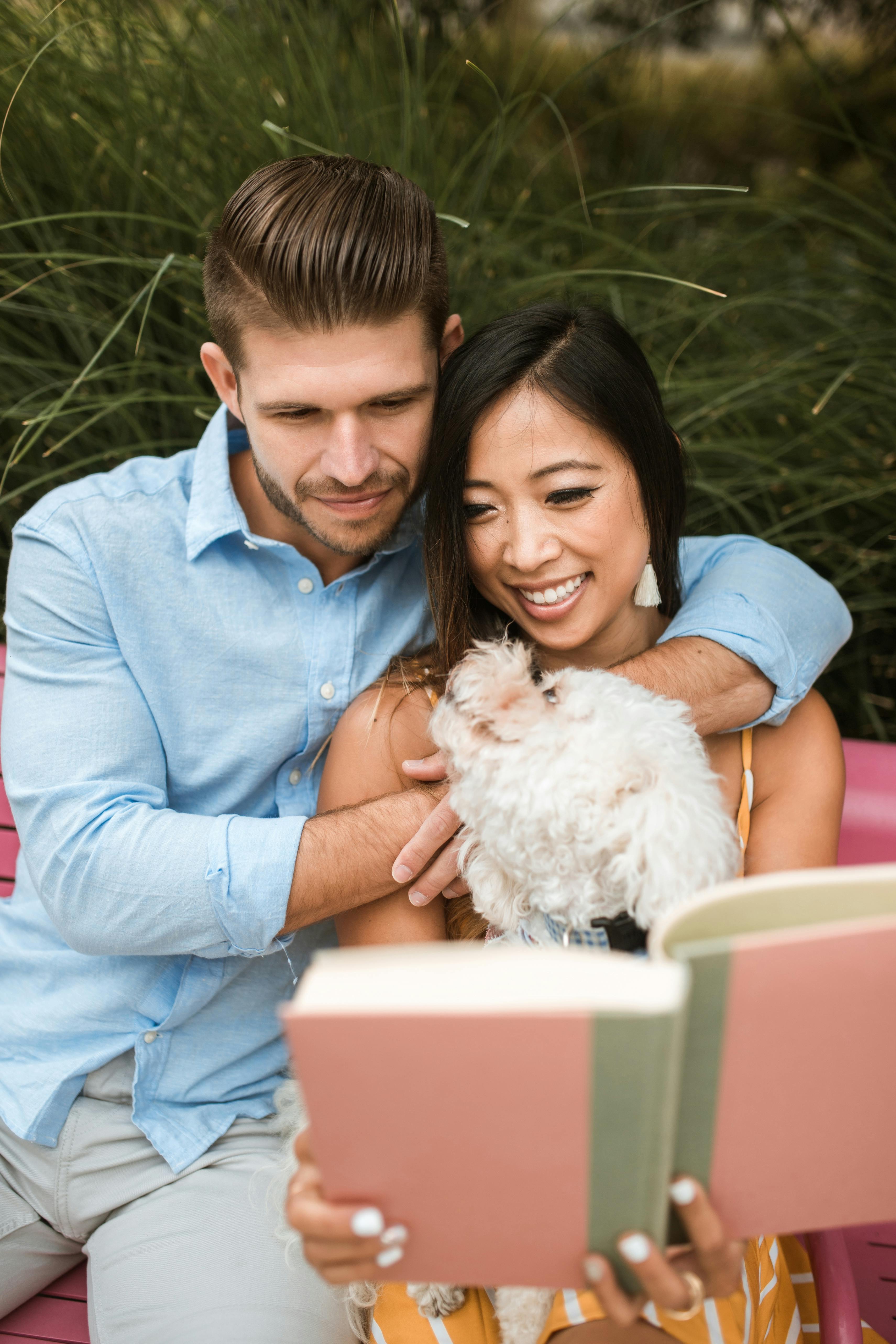 An affectionate couple enjoys reading a book outdoors with their Bichon Frise on a sunny day.