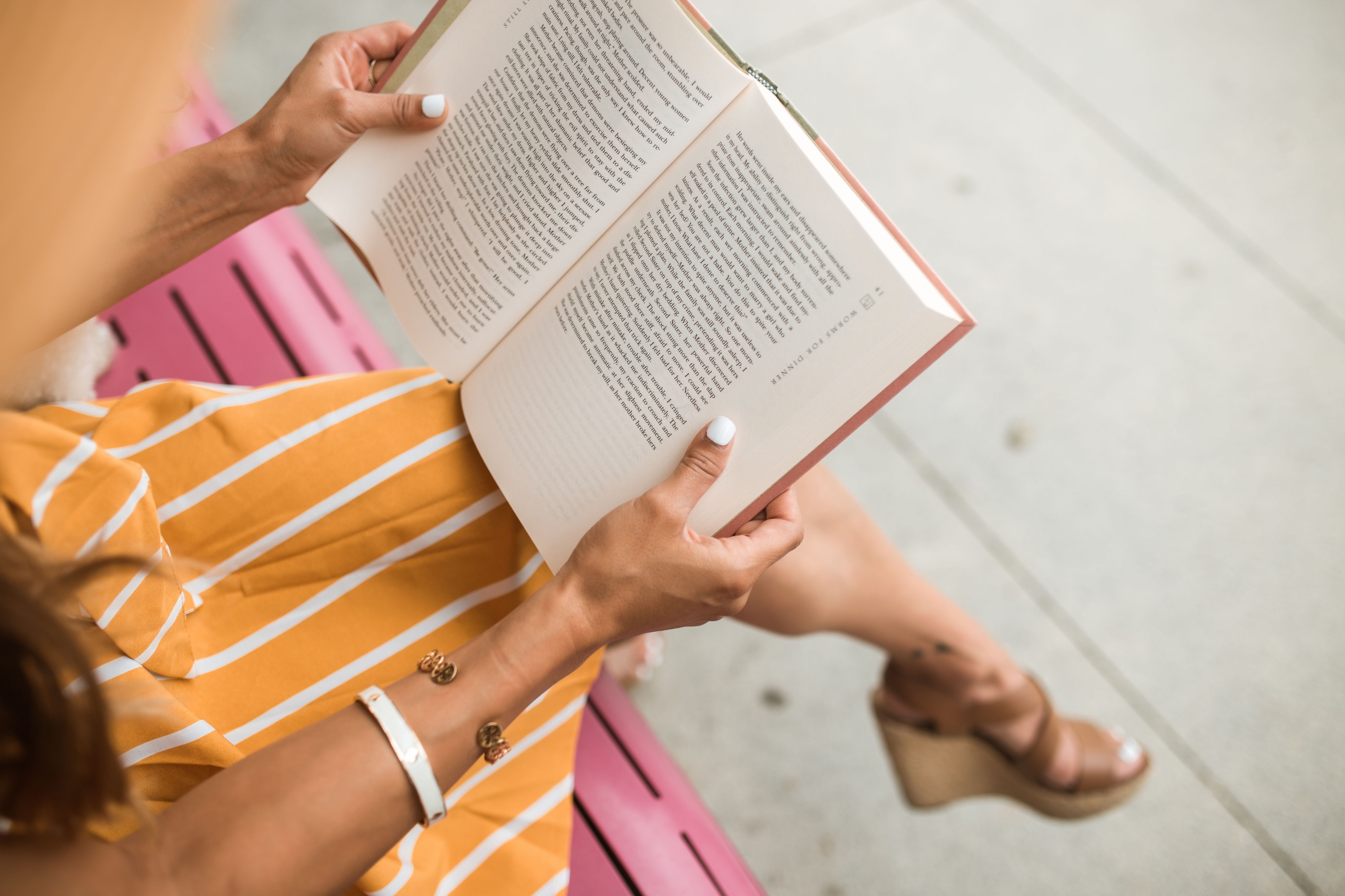 A woman in a striped dress reads a book while sitting on a pink bench outdoors.